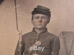 Civil War soldier holding rifle in front of a military camp backdrop tintype
